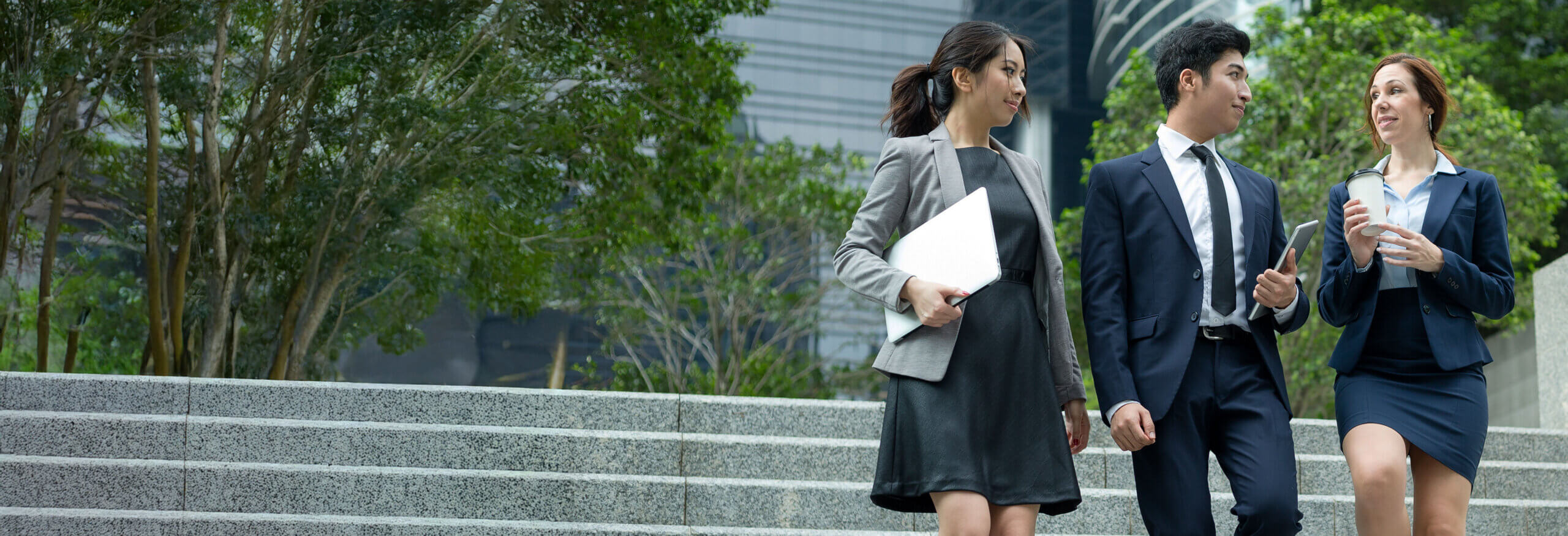 Three professionals in business attire walk up outdoor steps, carrying a laptop, tablet, and coffee cup.